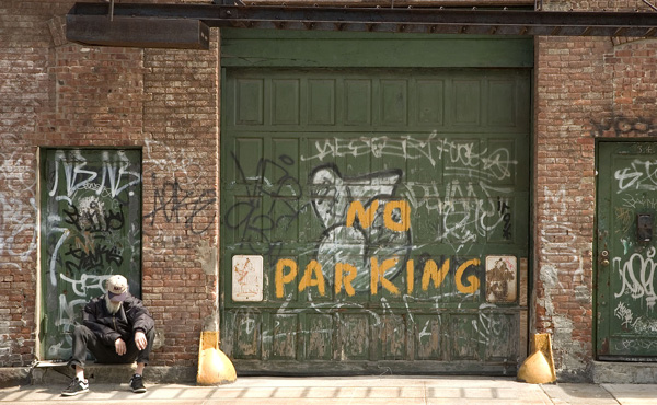 A homeless man sits on a stoop, next to a garage painted 'No
Parking.'