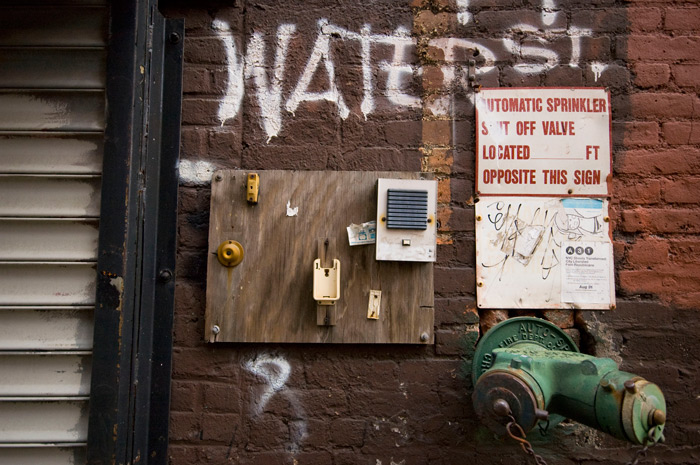 'Water Street' is spray-painted on a brick wall, which also has buzzers and a siamese water valve.