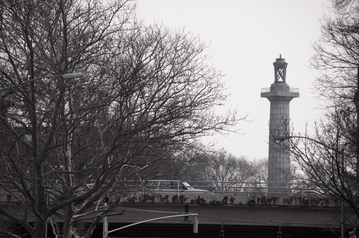 A tall column, topped by a bronze lantern, rises above the Brooklyn-Queens Expressway and surrounding trees.