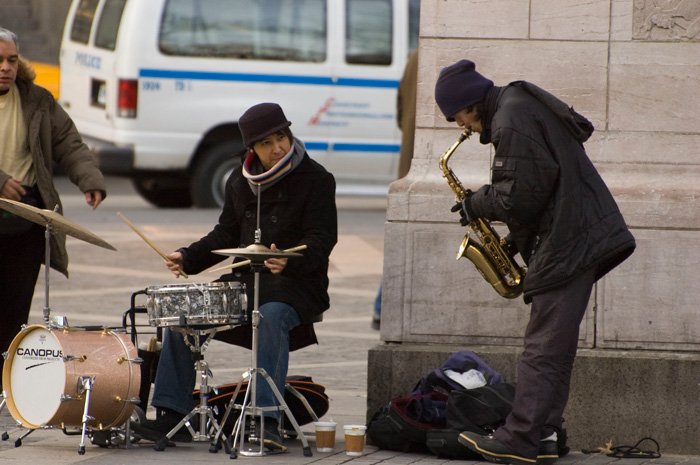 An alto sax player and a drummer, both dressed in black, play bebop outside an entrance to Central Park.