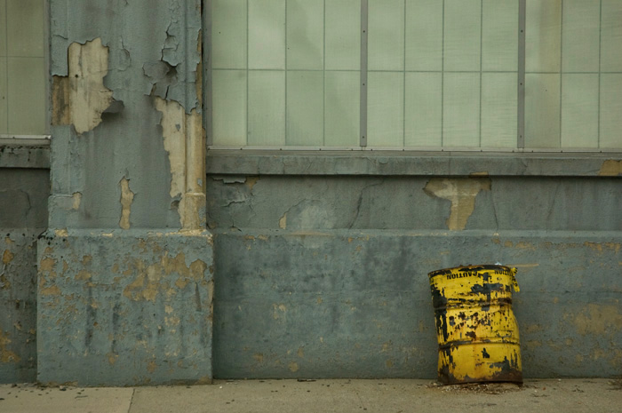 A battered, yellow, 50 gallon drum sits on the sidewalk, in front of a drab grey building.