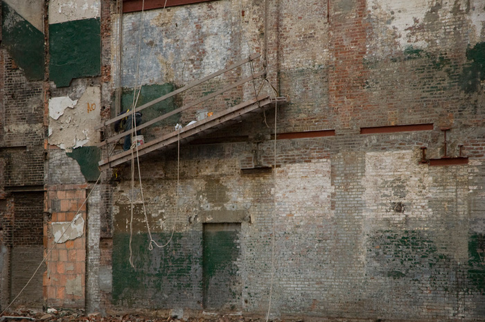 A scaffold tilts at about 45 degrees, as it hangs against a multi-colored exposed brick wall.