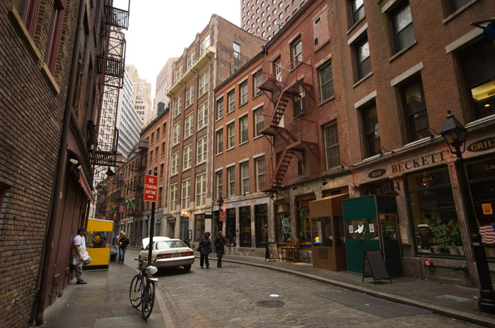 A winding street in Lower Manhattan is laid with Belgian paving stones and has mid-19th century buildings, but sky scrapers lay beyond its end.