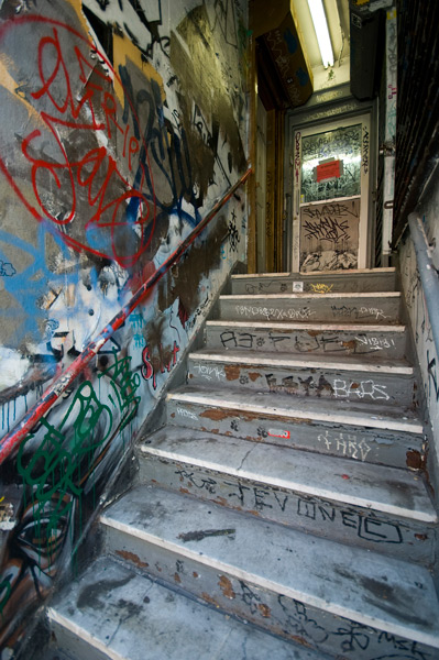 A stairway in a hall leads up to the parlor floor entrance of a building; the hall is covered in multi-colored paint and graffiti tags.