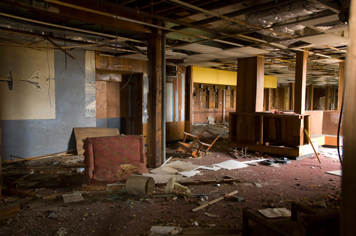 An abandoned room is littered with debris. Several chairs are overturned, and the ceiling is falling down.