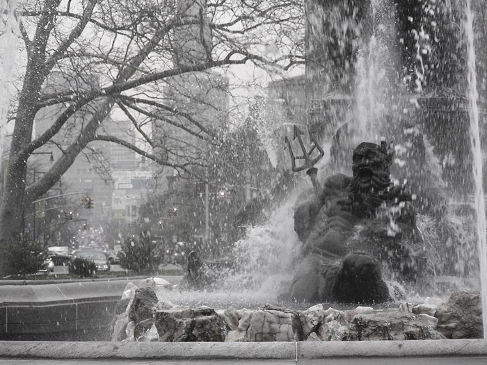 A figure of Neptune is showered by the waters of a water fountain, and city avenues stretch into the background.