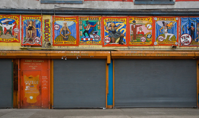 An orange door, with the word 'office' on it, sits beneath a row of colorful banners advertising members of a freak show.