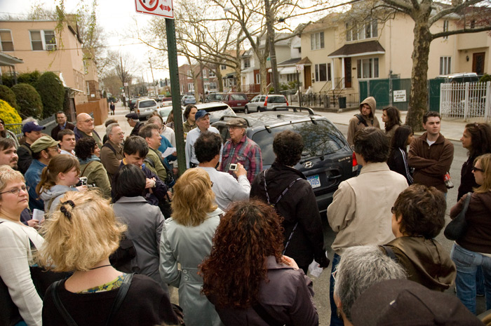 A group of attentive tourists on the sidewalk listen to a man describing his neighborhood.