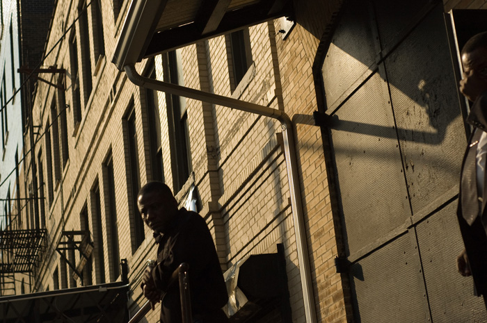 A man in silhouette leans against a railing in front of a brick building.