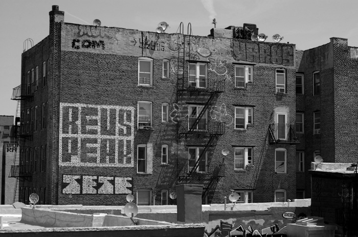 The street artist 'Revs' has painted his name in negative (holes where his letters are, on painted spaces) on the back wall of a large brick apartment building.