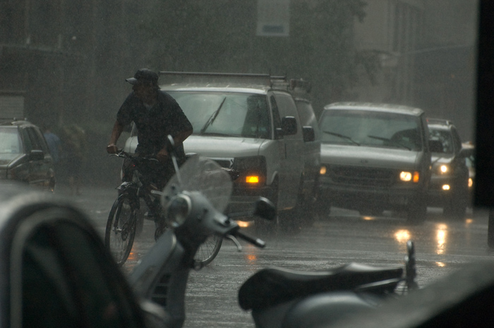 In a torrential downpour, a pizza delivery man pauses on his bicycle to judge traffic before turning.