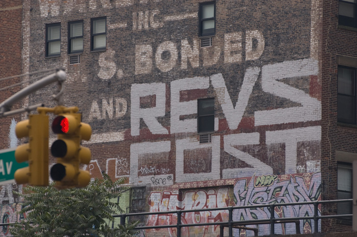 The side of a brick building has been painted with two large graffiti tags, for 'Revs' and 'Cost.' Painted with a roller, they maneuver around windows.