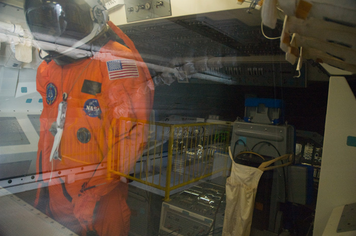 An orange flight suit, and helmet, stand behind the glass window of a display case within a space capsule.