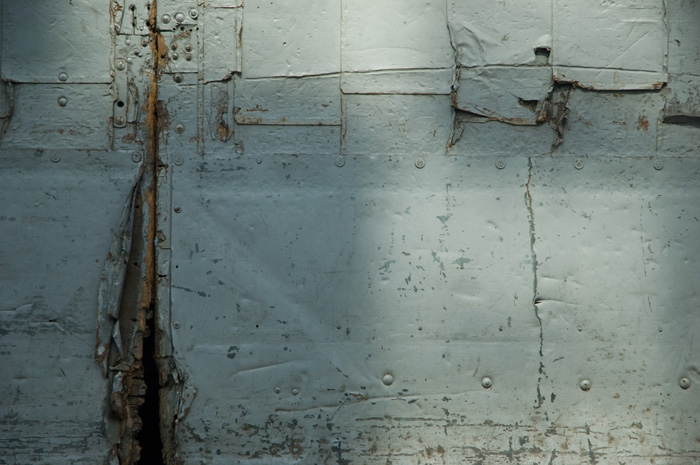 A weathered grey door shows craacks, peeling layers, and signs of several efforts to reinforce it.