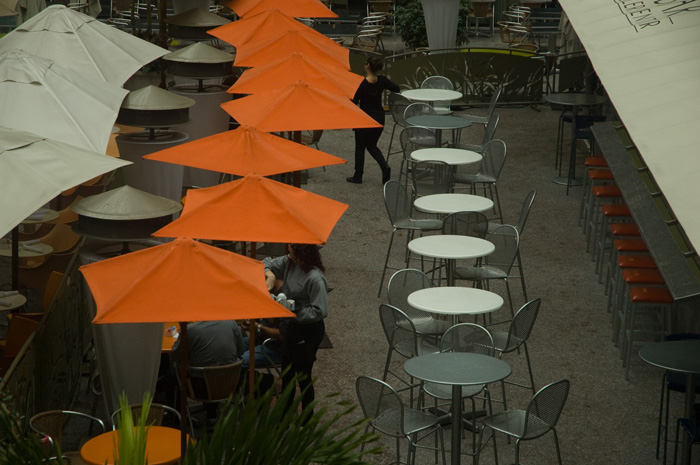Orange umbrellas shelter some diners, while tables without umbrellas are empty.