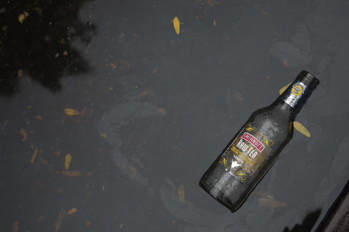 An empty bottle from a vodka beverage floats in a puddle of dirty, oily water.