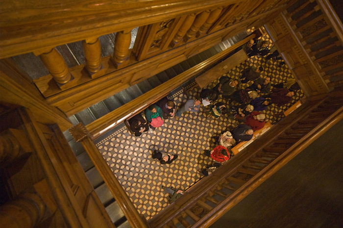 The picture looks down the middle of a wooden stairway, to a tour group and their guide, on a patterned tile floor.