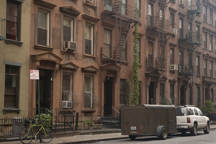 A sports vehicle and trailer are parked in front of a row of brownstones, one of which has a solitary vine running down the front.