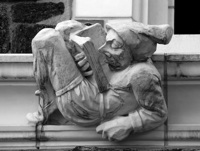 A building's 'gargoyle' is a figure in a medieval cap, lying on his back and concentrating on a book.