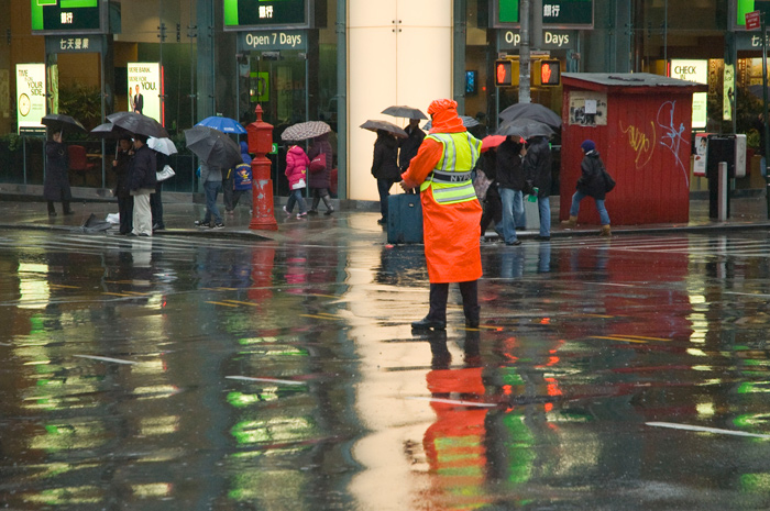 An orange-vested traffic cop stands in the middle of a rain-soaked intersection; reflections shimmer on the asphalt.