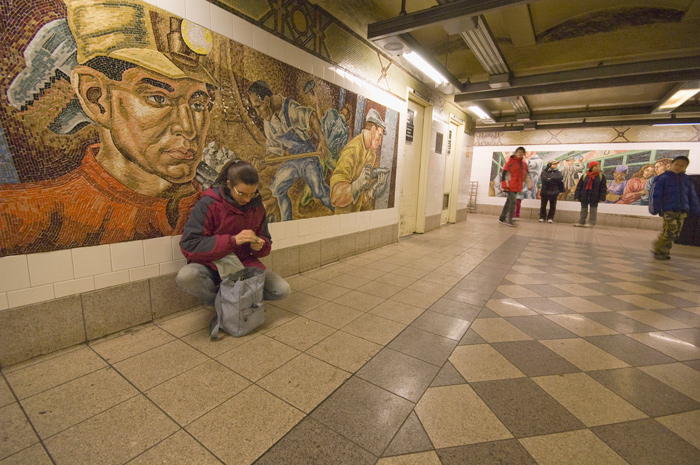 A photographer squats to change film, while in front of a large mosaic in a subway station.