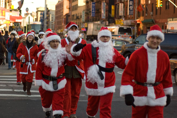 Several merry people in Santa Claus suits gesticulate as they cross a wide avenue.