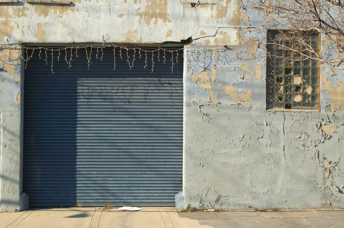 Icicle lights still hang from the wall around the garage door of an industrial building with grey peeling paint.