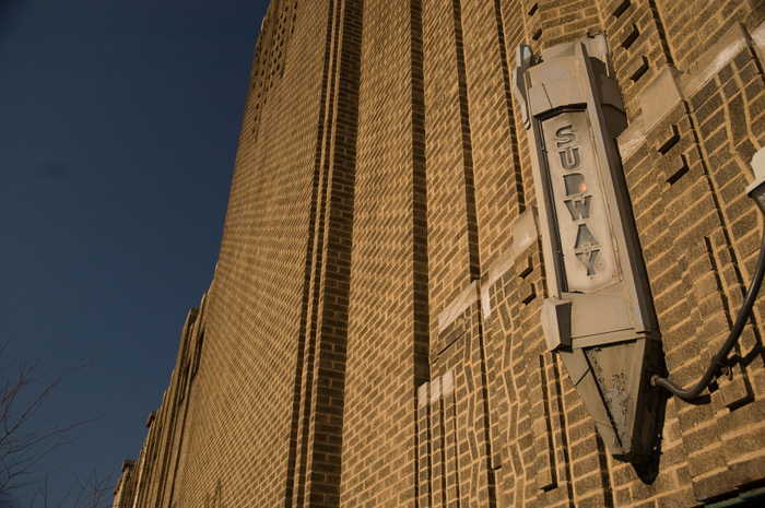 An art deco lighting fixture reads 'Subway,' attached to a large yellow brick structure.