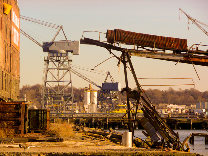 A chute descends from a trestle, but what fed the chute is gone. Shipping cranes are in the background.