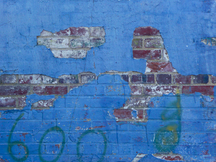 The layers of paint on a blue wall have been chipped away in an odd shape, showing the brick beneath.