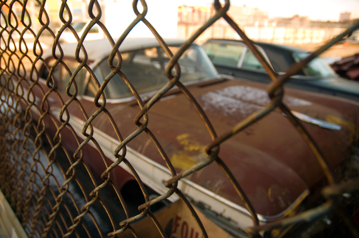 An old car is behind a chain link fence, with sunlight streaming beyond the car.