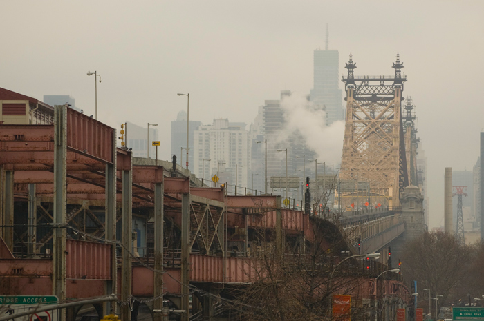 A roadway leads to the Queensborough Bridge, with steam rising in the distance, and Manhattan beyond that.