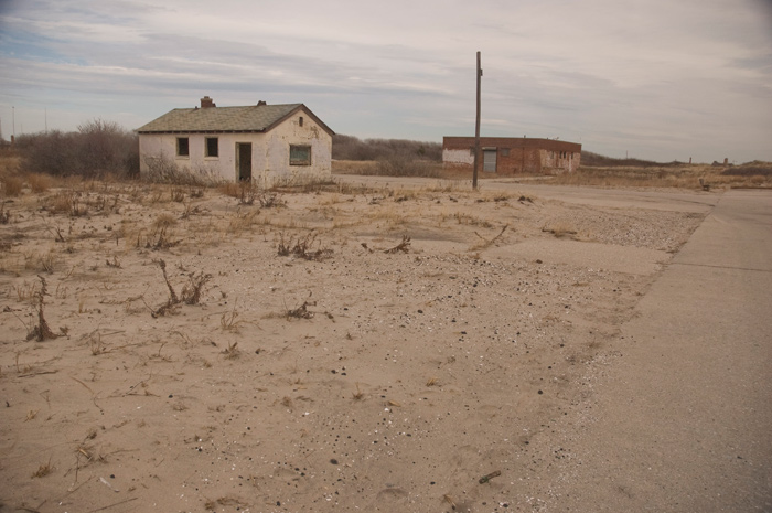 Two worn isolated buildings sit on sand and a concrete parking lot.