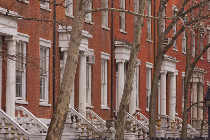 A series of brick row houses, with white entrances and columns, lies behind leafless trees.