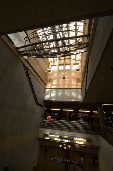 A sculpture suspended below a skylight casts shadows on stairs near a subway platform.