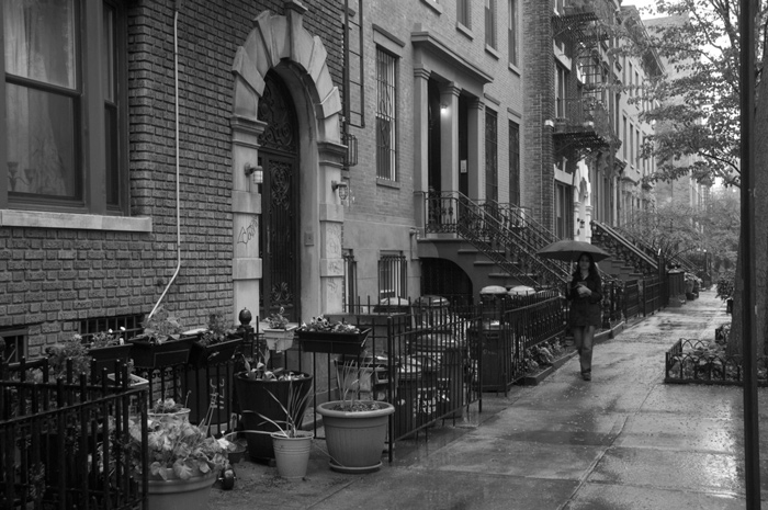 A woman with an umbrella walks past a row of brownstones on a rainy day.