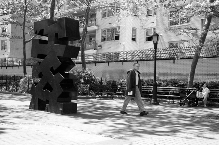 A man walks past a tall, black abstract sculpture in a tree-lined plaza.