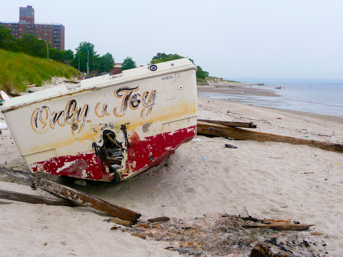 An old boat, with peeling paint, lies on the beach, with tall apartment buildings in the background.