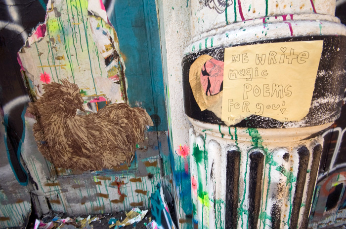 A wall and nearby pillar are decorated with posters and psychedelic paints, as well as a sign offering poetry for pay.