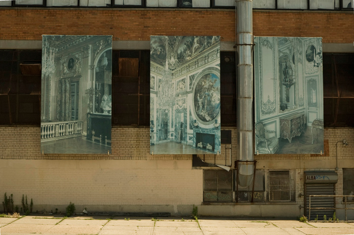 On the side of an industrial building hang three large, faded panels showing baroque furniture and room settings.