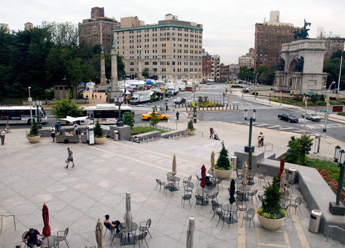 An arch with statuary at its top sits in the middle of a traffic circle; people sit at tables on a plaza.