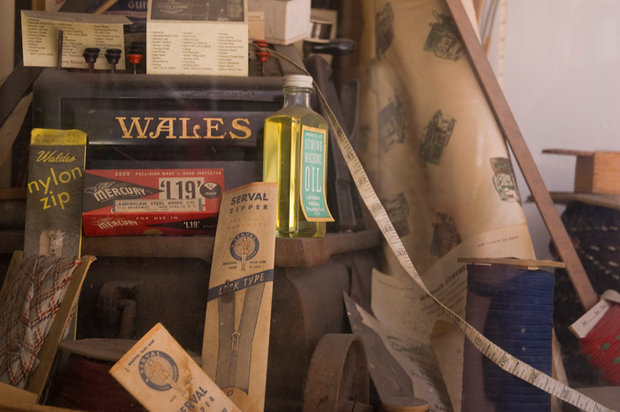 A shop window has old zippers, measuring tapes, fabrics, and sewing machine oil.