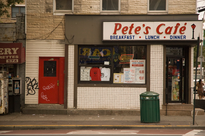 An external shot of a corner diner, decorated with white tiles and red and blue signage.