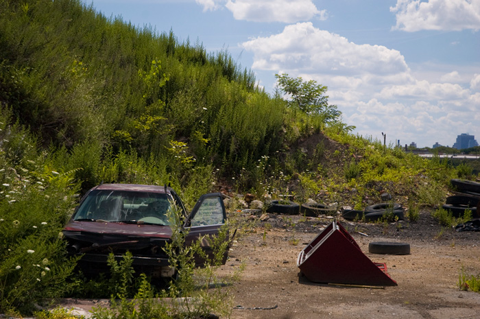 An abandoned car sits on a patch of dirt, next to a hill with tall weeds.