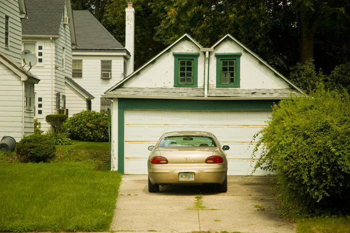 A compact car is parked on a concrete driveway, in front of a white and green, windowed garage.