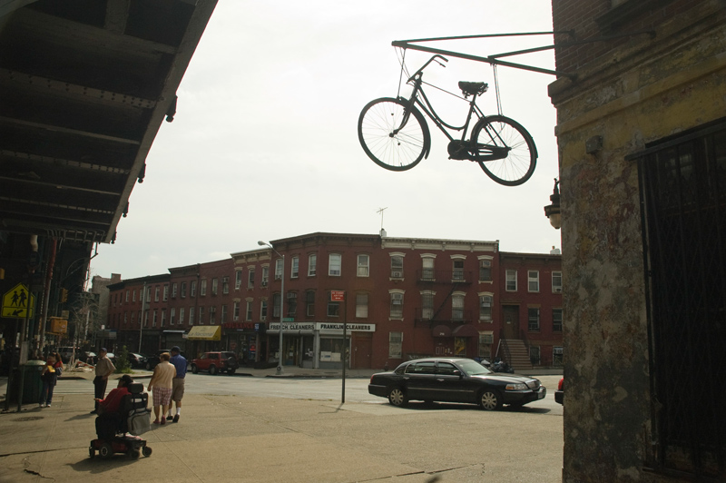 A bicycle hangs from the side of a building, and a handful of people cross a small triangle.
