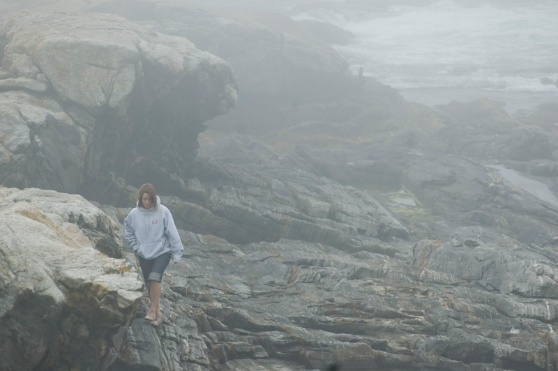 A tourist in jeans and hooded sweatshirt negotiates a rocky outcropping amidst fog from the sea.