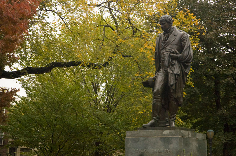 A statue of Robert Fulton, in a park with trees in Autumn colors.