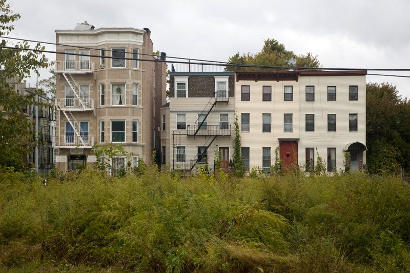 A string of row houses, with empty weed-filled lots in the foregeound.