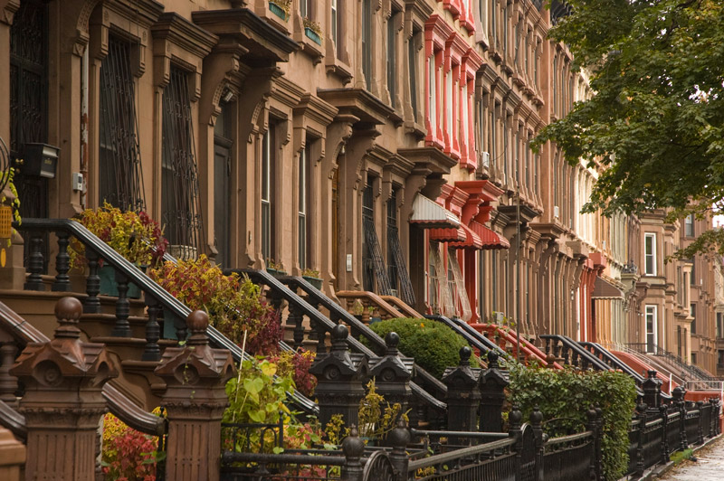 A street is lined with brownstone row houses and their stairs, each varying slightly from the other.